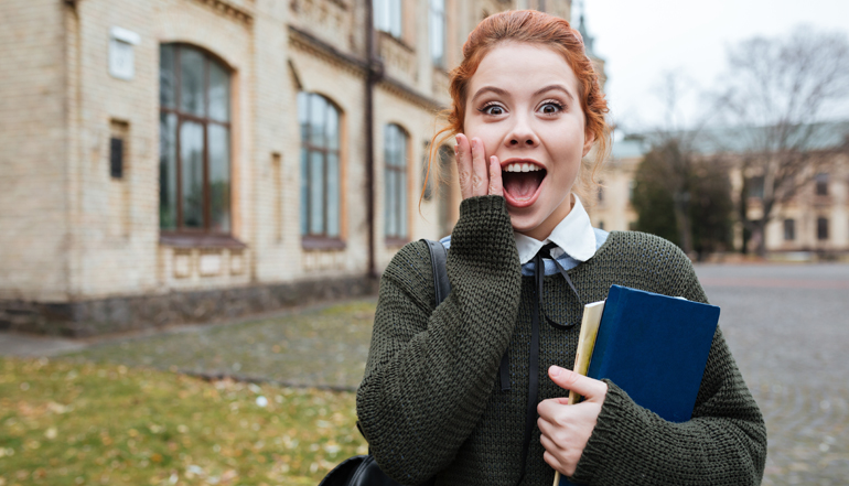 Astonished redheaded woman student holding books outside university campus