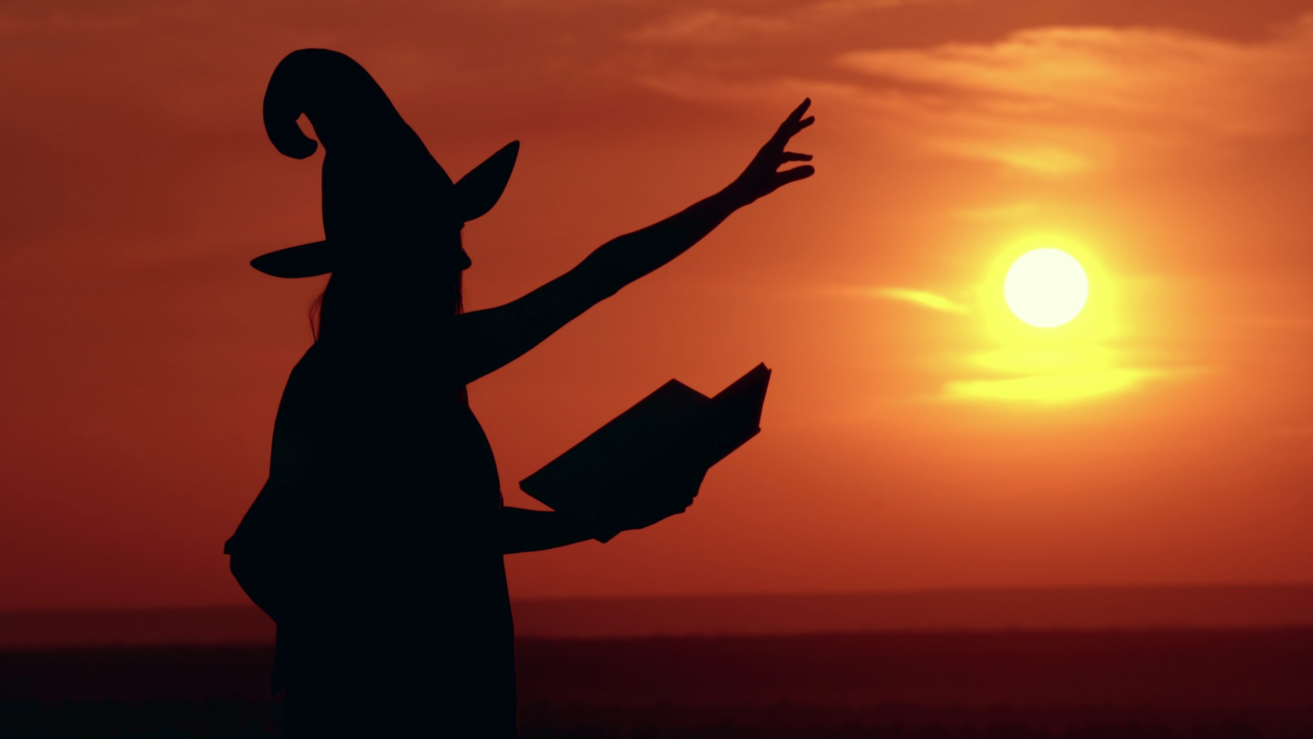woman-in-halloween-costume-sorceress-reading-spell-from-magic-book-on-the-hill-sky-wit-SBI-349426332